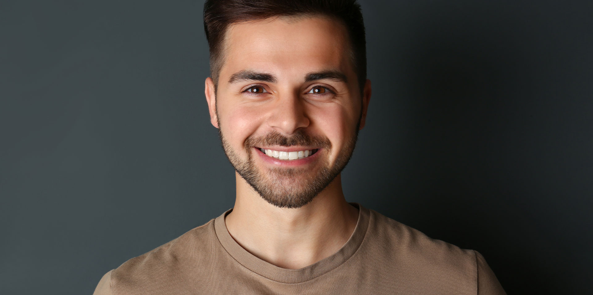 The image shows a smiling man with short hair, wearing a light-colored top, posing against a dark background.