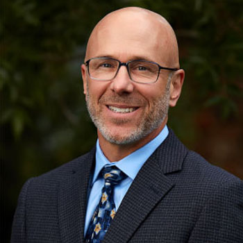 Man with bald head wearing glasses, smiling, in suit and tie, standing outdoors.