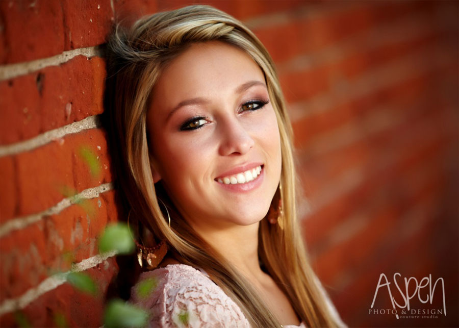 The image features a smiling woman with blonde hair leaning against a brick wall, wearing a pink top and earrings.